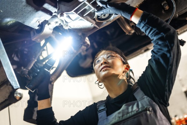 Young woman mechanic wearing safety glasses using flashlight while inspecting car chassis in a professional workshop