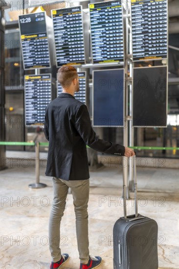Businessman with trolley suitcase looking at timetable screen while waiting his flight departure at the airport