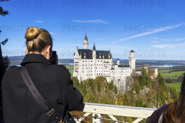 Woman photographing Neuschwanstein Castle in front of a picturesque landscape under a clear sky, Schwangau, Ostallgäu, Allgäu, Swabia, Upper Bavaria, Bavaria, Germany