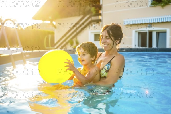 Mother and son having fun together in a home swimming pool, playing with a yellow inflatable ball on a sunny summer afternoon