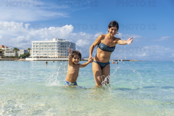 Mother and son holding hands and splashing in the crystal clear water of playa de s'estany d'en mas beach in mallorca, enjoying summer vacation