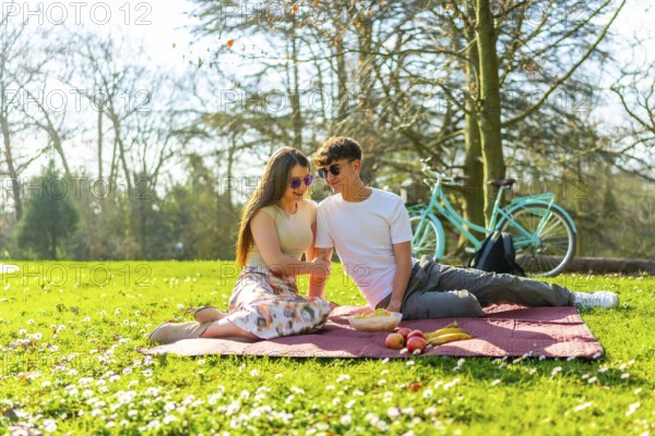 Happy couple sharing a romantic picnic in a sunny park with their bicycle nearby, enjoying fresh fruit and good company amidst blooming flowers