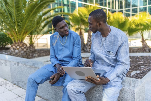 Two senegalese businessmen wearing traditional dashiki attire are discussing business while using a laptop outdoors