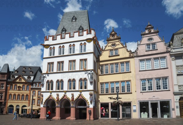 Colourful historic buildings in Gothic style with towers and windows on a lively town square under a blue sky, on the left Haus Steipe with arcade and battlements, Hauptmarkt, Trier, Rhineland-Palatinate, Germany