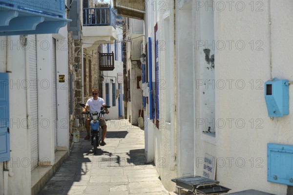 A narrow white alley with blue shutters. A man rides a motorbike through the sun, Mandraki, Nisyros, Dodecanese, Greek Islands, Greece