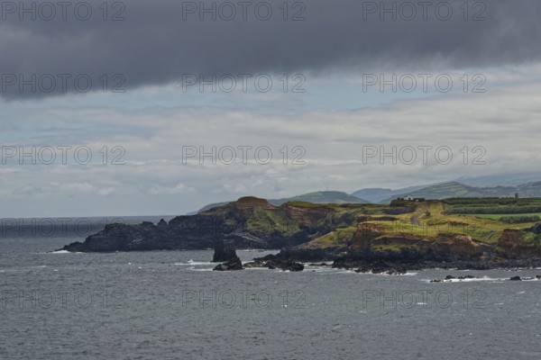A rocky coastline with green hills in the background under a cloudy sky, Fenais da Luz, Sao Miguel Island, Azores, Portugal