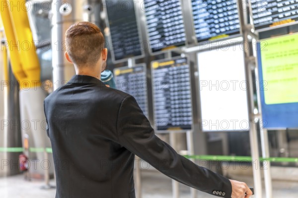 Businessman holding passport and pulling suitcase while checking flight schedule on departure board at airport terminal
