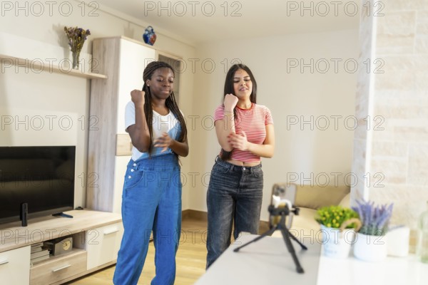 Two female friends dancing joyfully at home, capturing their fun moments on a smartphone while sharing their vibrant energy online