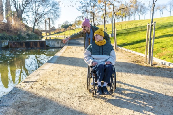 Man pushing friend in wheelchair, enjoying a sunny day in the park, surrounded by nature, trees, and a serene lake, sharing smiles and laughter