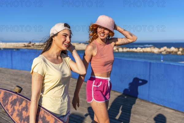 Two young women walk along a boardwalk by the sea, enjoying a sunny day. One carries a skateboard, both smiling and dressed casually