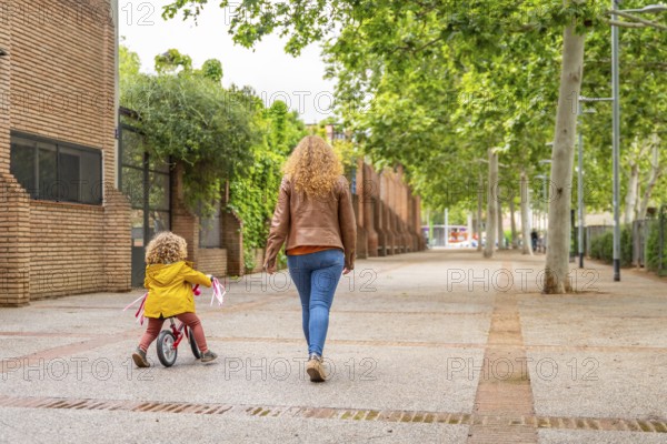 Rear view of a caucasian mother walking aside a girl learning to ride a bicycle in the street