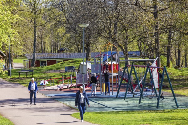 Playground in a public park with children playing and people walking on a footpath at springtime, Mösseberg, Falköping, Sweden