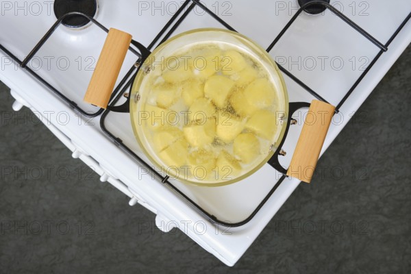 Sliced yellow potatoes are boiling in a clear glass pot on a white stove with wooden handles. The kitchen has a modern design, highlighting the process of preparing a meal