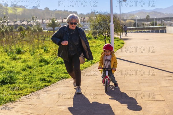 Grandmother jogging behind her grandson riding a balance bike in a park, having fun together on a sunny day