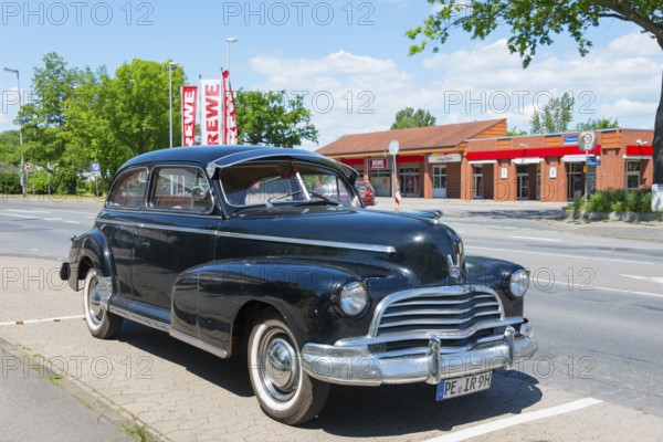 A black vintage car is parked in a car park next to a street with surrounding trees and shops, Chevrolet, Ilsede, Peine district, Lower Saxony, Germany