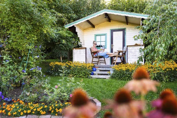 A woman sits relaxed on the small veranda of a garden shed and reads. Blooming summer flowers in the garden. Germany
