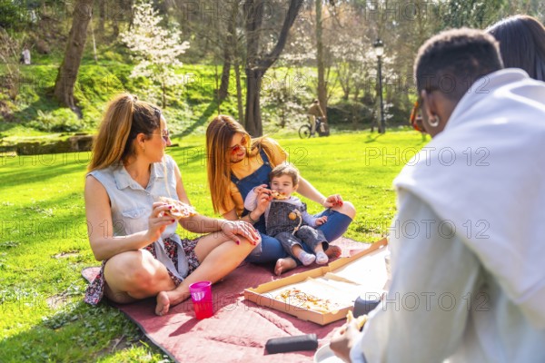Group of friends and family members are sitting on blanket, eating pizza and enjoying sunny spring day in park