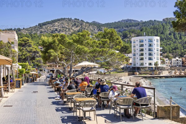 Outdoor restaurant with people on a pedestrian street in a coastal village by the beach with hotels at a tourist resort a sunny day, Sant Elm, Mallorca, Spain