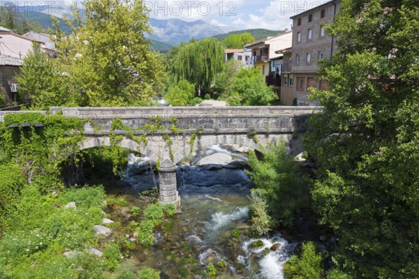 Eine historische Steinbrücke über einen klaren Fluss mit Blick auf Bäume, Gebäude und Berge im Hintergrund unter einem blauen Himmel, Arenas de San Pedro, Fluss Arenal, Ávila, Avila, Kastilien-León, Kastilien-Leon, Spanien