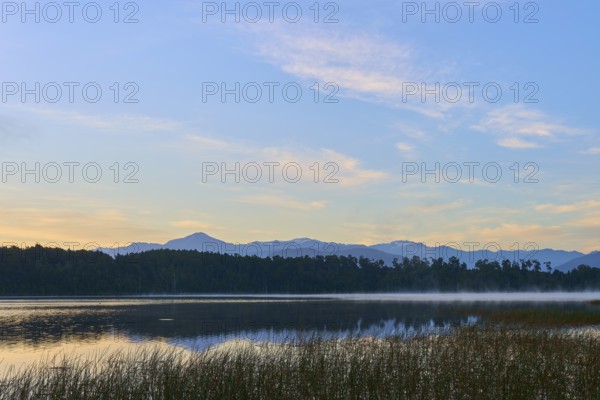 A calm, misty lake in morning sunlight with mountains in the background, Lake Mahinapua, Ruatapu, South Island, New Zealand