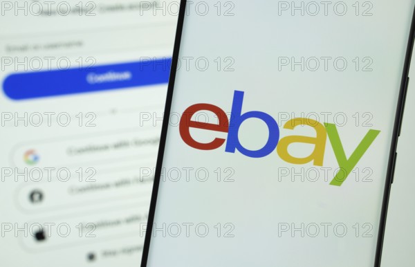 ebay logo is displayed on a smartphone screen, with the ebay login page visible on a computer screen in the background