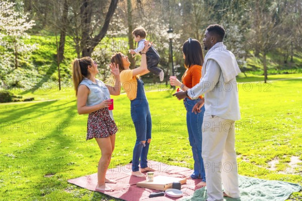 Cheerful multi ethnic family enjoying a sunny picnic on a green lawn in the park, happily lifting their baby and sharing joyful moments together