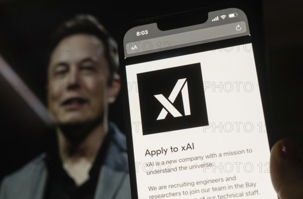In this photo illustration, A person browses website of xAI, displayed on a mobile phone, Elon Musk images in the background screen, in Guwahati, India 0n 15 July 2023. Tesla CEO Elon Musk launches Artificial Intelligence company xAI on 12 July 2023, the company's goal is to understand the true nature of the universe