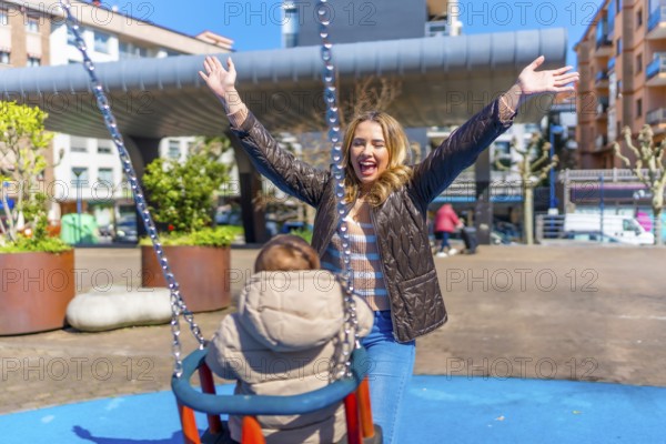 Young mother having fun pushing her baby on a swing in an urban playground, enjoying a sunny day outdoors