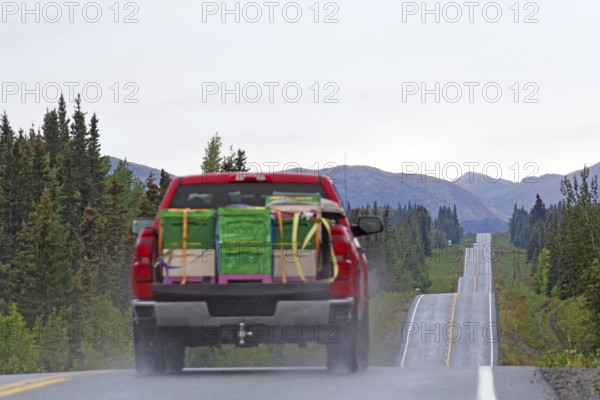 Red lorry with bulky goods drives up and down a straight road lined with forests and mountains, Richardson Highway, Alaska, USA