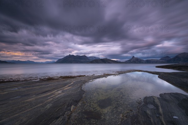 The Vestfjord in Nordland, Norway, offers a dramatic atmosphere near Bodø. Dark clouds are illuminated by the setting sun in shades of red and yellow. The rugged cliffs and mountains are reflected in the quiet fjord, which offers a breathtaking view of Lofoten