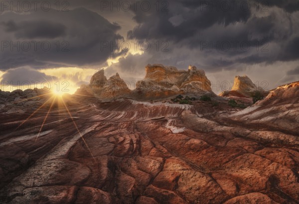 A dramatic sunset casts golden light over a rugged desert landscape, highlighting the textures of the red rock formations beneath a moody, cloud-filled sky