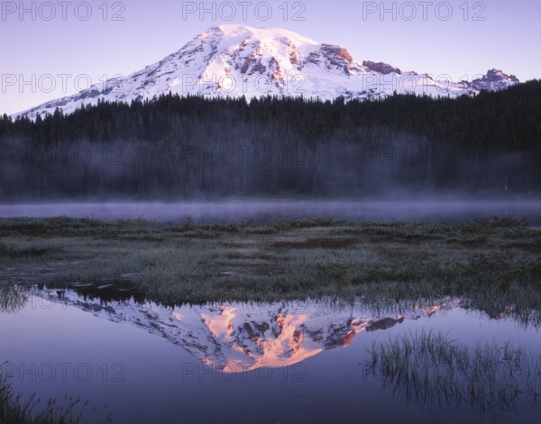Snow capped mountain reflecting in a calm lake at dusk, surrounded by mist and lush forest. The sun graces the peak, creating an enchanting and serene landscape