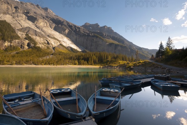 A serene view of Oachinensee in Switzerland captures several blue boats docked on the calm lake, reflecting the surrounding majestic mountains and vibrant green forests