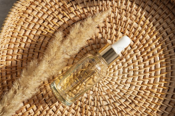 A glass cosmetic with golden essential oil rests on a woven basket alongside dried grass. The natural setting highlights self care and wellness concepts