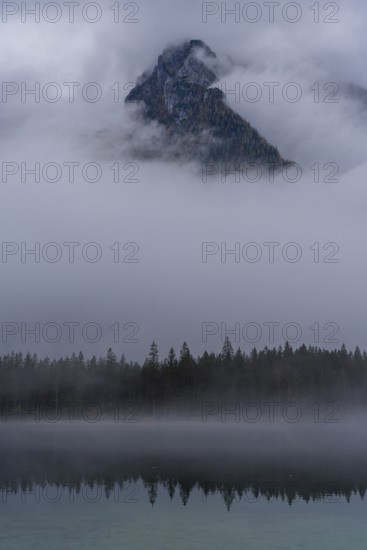 A stunning view of a mountain peak in Hintersee, partially shrouded in fog, surrounded by serene forest and reflected in a tranquil lake, capturing the ethereal beauty of the Bavarian Alps