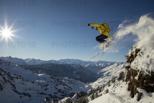 A skier in a yellow jacket performs an exhilarating jump off a snowy cliff, surrounded by a breathtaking mountain landscape under a clear blue sky and bright sun