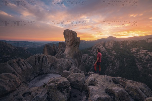 Back view of unrecognizable man standing on rocky outcrop, gazing at vibrant sunset over Pedriza Caballeros. Majestic rock formations and expansive views of Sierra de Guadarrama