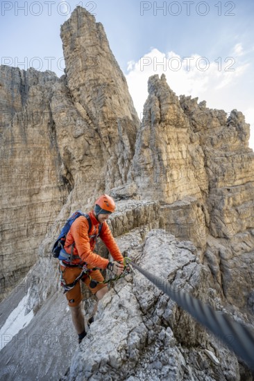 Mountaineer climbs an exposed rock in the secured Via Ferrata Bocciere Centrale via ferrata, spectacular rock towers in the back, Brenta Mountains, Brenta-Adamello Natural Park, Trentino, Italy