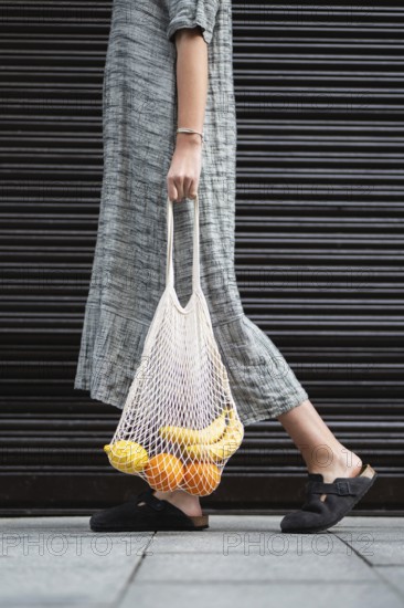 A woman in a long, gray dress carries a reusable mesh bag filled with bananas and oranges. The individual walks on a sidewalk, wearing black slip-on shoes