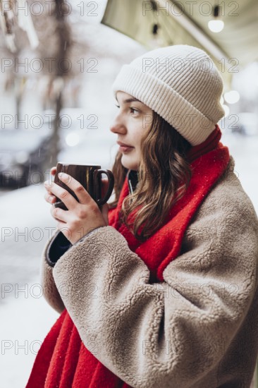 A young thoughtful woman in a white beanie and red scarf holds a warm mug, gazing thoughtfully while surrounded by a snowy ambiance