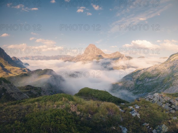 Serene mountain peak rises above a lush green landscape enveloped in wispy clouds under a vast blue sky in summer. The scene captures the essence of tranquility and natural beauty in the Pyrenees