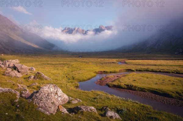 A serene mountain valley landscape with a winding river and mist shrouded peaks in summer. The warm sunlight gently illuminates the lush green terrain and rocky foreground in the Pyrenees