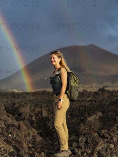 A woman stands amidst volcanic terrain at Timanfaya National Park, Lanzarote, enjoying a vibrant rainbow backdrop. Her smile reflects the wonder of nature's beauty