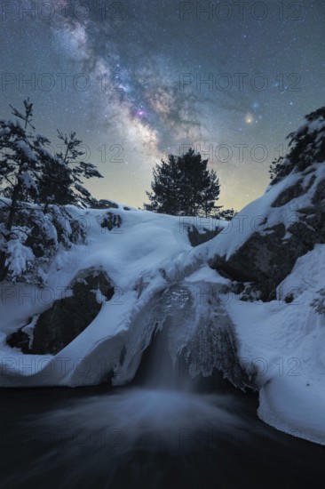 Captivating night scene depicting the Milky Way over a snow-laden landscape in Iceland A frozen waterfall adds an enchanting touch to this serene winter wonderland