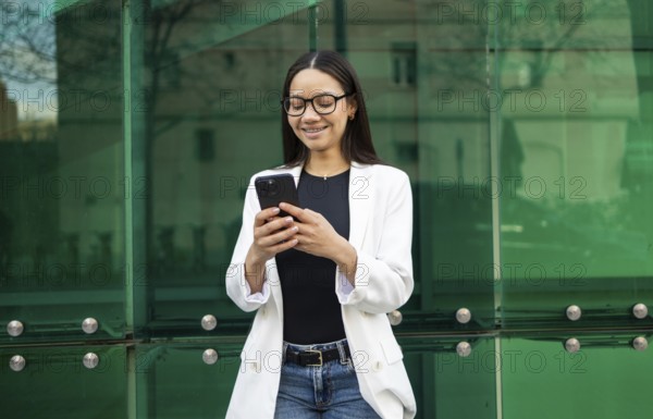 Hispanic businesswoman wearing glasses and a white blazer, smiling while using her smartphone against a modern glass building background. Urban business lifestyle scene