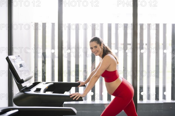 Pregnant woman in a red sports outfit exercising on a treadmill, promoting pregnancy wellness and active lifestyle. Bright, modern gym environment with large windows