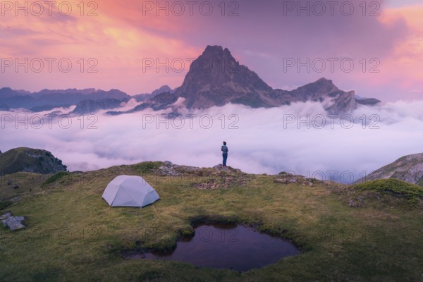 A camper stands near a tent overlooking a majestic mountain peak above a sea of clouds during a vibrant sunrise in the Pyrenees. The scene is peaceful and inspiring in summer