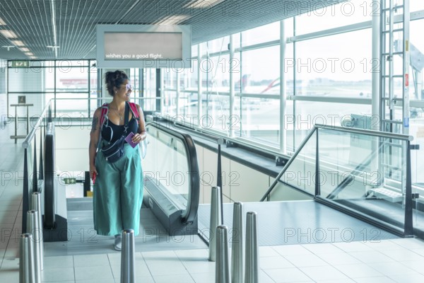 A traveler stands on an escalator in a bright airport, holding a passport, A sign above wishes you a safe flight Glass walls reveal airplanes outside