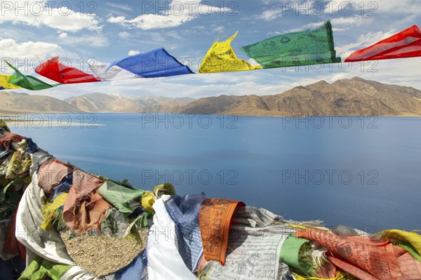 Colorful prayer flags flutter against the backdrop of the serene Pangong Tso lake, set amidst the rugged mountains of Ladakh, India, under a vibrant blue sky