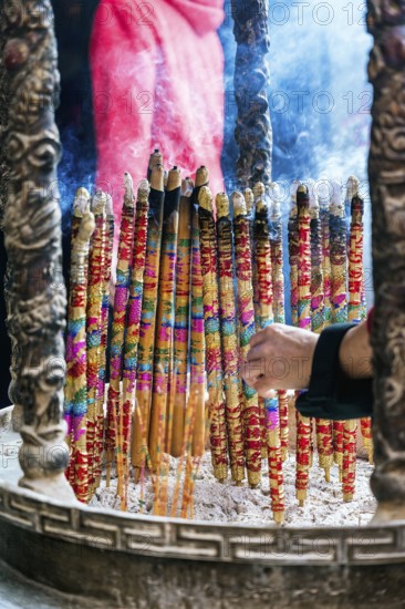 Cropped unrecognizable person holding a vibrant, large incense sticks burning, with smoke rising in a Taoist and Chinese Buddhist temple in Macau. A person is visible in the background adjusting the sticks